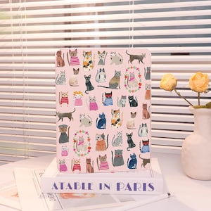May include: A pink tablet case featuring a repeating pattern of various cat illustrations. The cats are depicted in different poses and outfits, with a few surrounded by floral wreaths. The case is resting on a stack of books with the text "ATABLE IN PARIS".