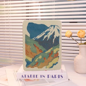 May include: A rectangular tablet case featuring a stylized mountain landscape print with a snow-capped peak in dark blue, white, and earthy green tones. The case rests on a white book with the text ATABLE IN PARIS printed in blue letters.
