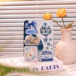 Könnte beinhalten: Ein Notizbuch mit Aufklebern von Schloss, Büchern und Sternen auf blau-weiß gestreiftem Hintergrund. Das Notizbuch hat eine blaue Schleife und die Aufschriften "Fantasy Book Club" und "Bookish Girlie". Daneben steht eine Vase mit gelben Blumen.