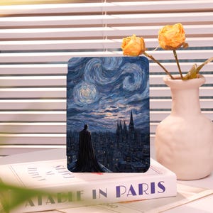 May include: A rectangular case featuring a Batman figure overlooking a cityscape under a starry night sky, reminiscent of Van Gogh's style. The case is resting on a book titled "A Table in Paris".