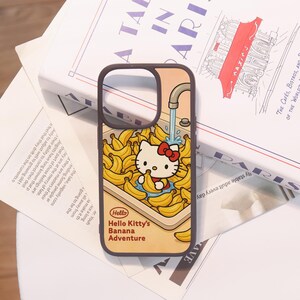 Może przedstawiać: Brązowe etui na telefon z motywem Hello Kitty w bananach. Etui przedstawia Hello Kitty trzymającą banana pod kranem, w otoczeniu banan&oacute;w. Widoczny jest r&oacute;wnież tekst "Hello Hello Kitty's Banana Adventure".