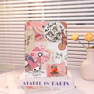 May include: A decorative case with a collage design, featuring illustrations and text. The design includes a My Melody character, a Hello Kitty CD, and a package of Big Cherry candy. The text includes "The Good, The Bad, The Average" and Japanese characters.
