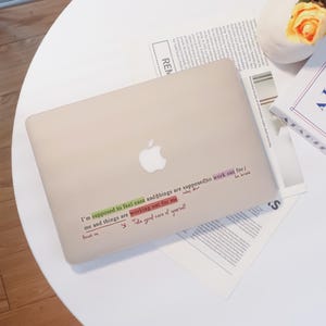 Könnte beinhalten: Ein beiges Laptop mit dem Apple-Logo liegt auf einem weißen Tisch. Auf dem Laptop befindet sich ein Aufkleber mit mehrfarbigem Text. Der Text lautet: "I'm supposed to feel ease and things are supposed to work out for/ be brave me and things are working out for me. focus on Take good care of yourself."