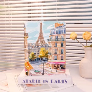 May include: A rectangular art print depicting a Parisian street scene with the Eiffel Tower. The print is placed on a book with the text "ATABLE IN PARIS". A vase with yellow flowers is to the right. The print shows buildings and a car.