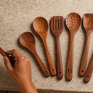 May include: A selection of wooden kitchen utensils, including spoons, spatulas, and slotted tools. The utensils are made of a warm-toned wood and are arranged on a light-coloured countertop. One utensil is being held over a pan.