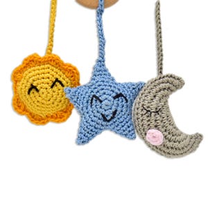 May include: A handmade baby mobile featuring a yellow sun, a blue star, and a gray crescent moon, all crocheted with smiling faces. The sun has orange rays, and the moon has a pink cheek. The mobile is suspended from a wooden ring.