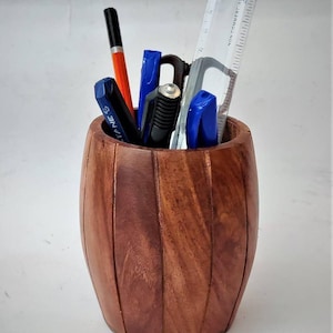May include: A wooden barrel-shaped desk organizer, holding pens, a ruler, and other stationery items. The organizer is made of dark brown wood and has a natural grain. The pens are various colors, including black, blue, and orange.