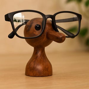 May include: A wooden eyeglass holder shaped like a whimsical character with a long nose and a black eye. Black-framed eyeglasses rest on the holder. The holder is made of dark brown wood and sits on a light-colored surface.