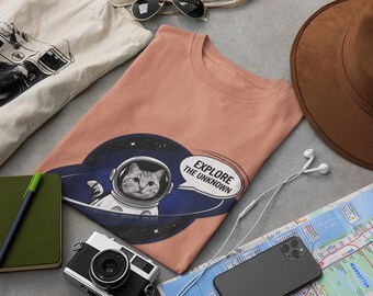 Könnte beinhalten: Ein pfirsichfarbenes T-Shirt mit einem Aufdruck eines Katers, der einen Astronautenhelm trägt, und einer Sprechblase mit dem Text "Explore the Unknown".