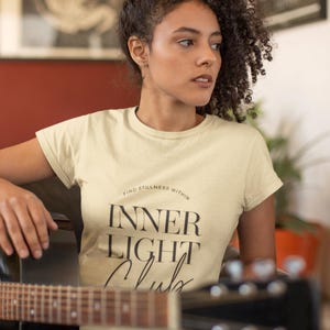 May include: A woman wearing a light yellow t-shirt with the text "Find stillness within Inner Light Club" printed on it. She is holding a black acoustic guitar.