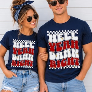 May include: Two navy blue short-sleeve t-shirts featuring a graphic print with a black and white checkered border. The text reads HELL YEAH DAMN RIGHT in a wavy, retro-style font with alternating white and red letters and small gold star accents.