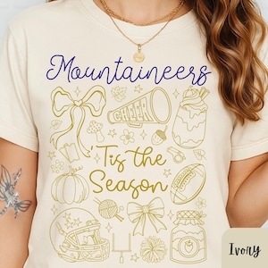 May include: Ivory-colored t-shirt with the words "Mountaineers" in blue script and "'Tis the Season" in gold script. The shirt features gold line art of a megaphone, football, and other seasonal and cheerleading-themed graphics.