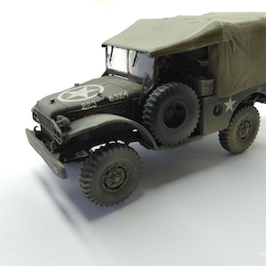 May include: An olive drab military truck model featuring a canvas-covered cargo bed, a spare tire mounted on the side, and a white star insignia on the hood. The hood displays the numbers 223 and 8324, and the vehicle has rugged treaded tires.