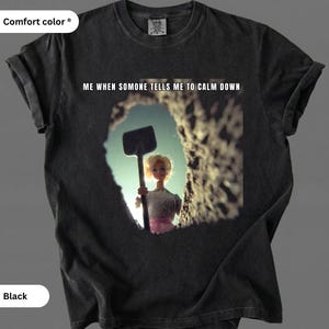 May include: Black Comfort Colors t-shirt featuring the text "ME WHEN SOMEONE TELLS ME TO CALM DOWN." The graphic shows a doll holding a shovel in a hole. The shirt is a dark, solid colour.
