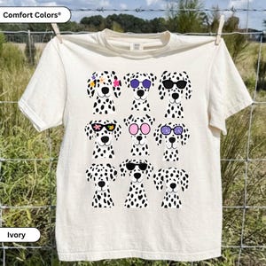 May include: An ivory short-sleeved t-shirt featuring a graphic print of nine dalmatian dog heads arranged in a grid. The dogs are wearing various styles of colourful sunglasses and floral accessories. The shirt has a crew neckline and short sleeves.