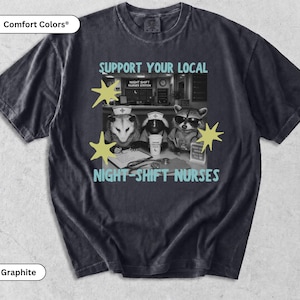 May include: A graphite-colored t-shirt featuring a graphic of three animals dressed as nurses at a station. The text reads SUPPORT YOUR LOCAL NIGHT-SHIFT NURSES. The animals include an opossum, a skunk, and a raccoon wearing nurse caps.