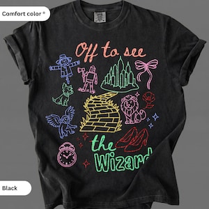 Op de afbeelding: Zwart t-shirt met een kleurrijk grafisch ontwerp met personages en elementen uit The Wizard of Oz. De zin "Off to see the Wizard" is geschreven in een grillig lettertype. Het shirt is gemaakt van Comfort Color materiaal.