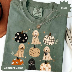 Può includere: Una t-shirt a maniche corte color verde muschio con una stampa grafica di tre cani doodle chiari, un cane doodle nero e cinque zucche con vari motivi tra cui margherite, quadri e pois. La maglietta presenta un'etichetta Comfort Colors.