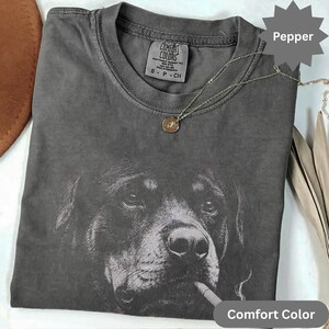 Könnte beinhalten: Ein anthrazitfarbenes T-Shirt mit Rundhalsausschnitt und einem Grafikdruck des Gesichts eines Rottweilers. Das T-Shirt hat ein Comfort Colors Markenetikett am Innenkragen mit dem Text S P CH. Eine dünne goldene Kette liegt auf dem Stoff.