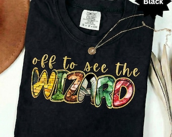 Op de afbeelding: Een zwart t-shirt met korte mouwen en een ronde hals. Het shirt is voorzien van de tekst "off to see the WIZARD" in gouden letters. De letters van het woord WIZARD bevatten grafische illustraties van een gele bakstenen weg, een leeuw en bladeren.