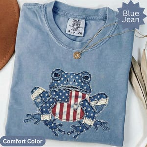 May include: Blue jean Comfort Colors t-shirt with a frog graphic. The frog is made of denim with a patriotic design, featuring stars and stripes. A gold necklace with a pendant is draped across the shirt.