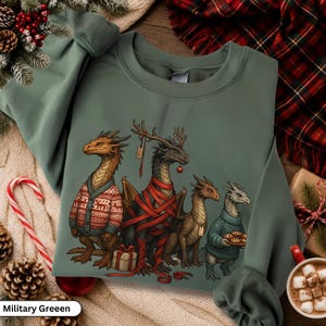 Puede incluir: Una sudadera verde militar con un gráfico de cuatro dragones. Un dragón lleva un cárdigan con estampado rojo y blanco, otro está envuelto en cinta roja con un regalo, uno tiene astas y otro lleva un suéter azul sosteniendo un plato de galletas.