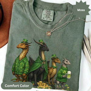 May include: A moss green Comfort Colors t-shirt featuring a St. Patrick's Day dragon design. The graphic includes four dragons wearing green hats and sweaters. A silver necklace with a pendant is also visible.