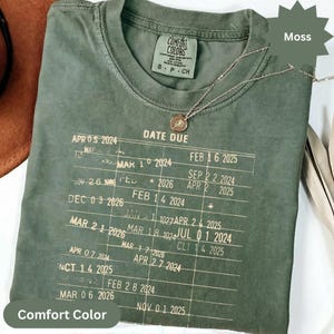 May include: A moss green Comfort Colors t-shirt with a printed chart of dates and the words "DATE DUE". A gold-colored necklace with a pendant rests on the shirt. The shirt is folded.
