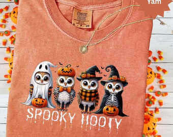 May include: A Comfort Colors orange-colored t-shirt with a crew neck. The shirt features a graphic of four Halloween-themed owls with pumpkins and the text "Spooky Hooty". A gold necklace with a pendant is draped on the shirt.