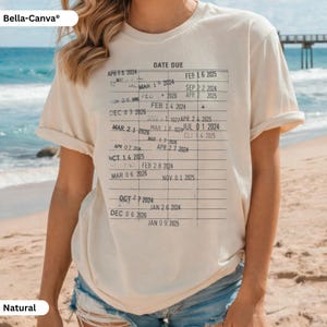 May include: A natural-colored t-shirt featuring a printed library card design with the words "DATE DUE" at the top. The design includes a grid of dates and lines, creating a vintage aesthetic. The shirt is from Bella+Canvas.
