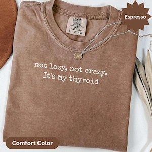 May include: An espresso-colored short-sleeve t-shirt with a crew neckline. The shirt features white printed text that reads, "not lazy, not crazy. It's my thyroid." A silver-toned chain necklace with a small circular pendant rests on the collar.