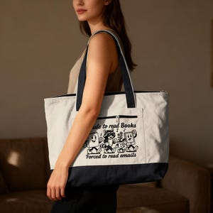 Peut inclure: Un sac fourre-tout blanc avec un fond et des sangles bleu marine. Le sac présente un dessin animé en noir et blanc avec le texte "Made to read Books, Forced to read emails". Il a un dessus zippé et une poche avant.