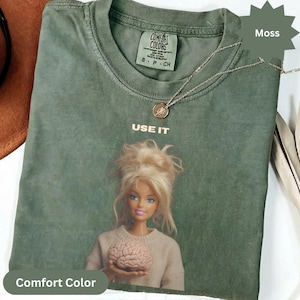 Può includere: Una t-shirt a maniche corte color verde muschio con la grafica di una bambola bionda che tiene in mano un cervello. Il testo "USE IT" è stampato sopra la bambola. La maglietta ha un'etichetta Comfort Colors sul colletto e una collana dorata.