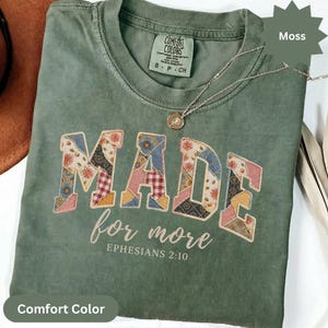 May include: A moss green t-shirt featuring the word MADE in large block letters with a multi-coloured patchwork floral and gingham pattern. Below, the text reads for more EPHESIANS 2:10 in a white script and block font. A thin gold necklace rests on the collar.