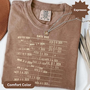 May include: Espresso-colored Comfort Colors t-shirt with a printed "Date Due" design. The shirt features a crew neck and short sleeves. A silver necklace with a pendant is draped on the shirt. The shirt is labeled "Comfort Colors" and the color is "Espresso".