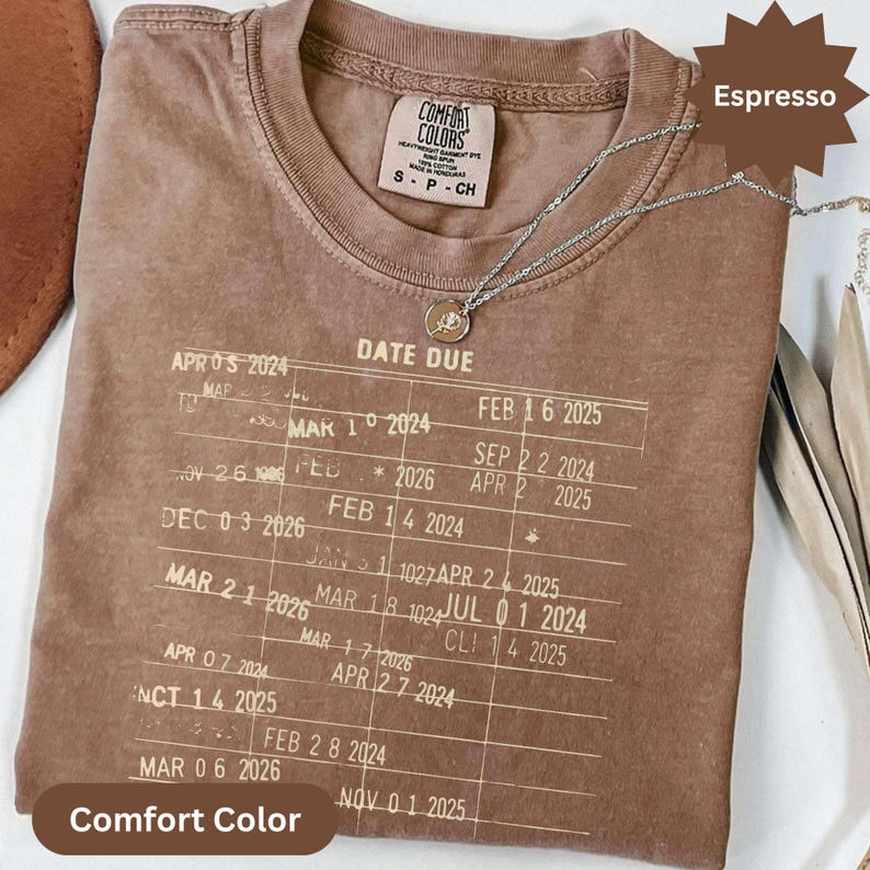 May include: Espresso-colored Comfort Colors t-shirt with a printed "Date Due" design. The shirt features a crew neck and short sleeves. A silver necklace with a pendant is draped on the shirt. The shirt is labeled "Comfort Colors" and the color is "Espresso".