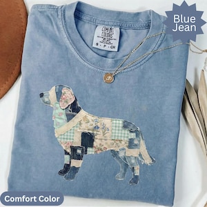 Op de afbeelding: Een blauw spijkerkleurig t-shirt met een patchwork hondengrafiek gemaakt van diverse bloemen- en ruitpatronen. Het shirt heeft een ronde hals en een Comfort Colors label in de kraag. Een dunne gouden ketting ligt op de stof.