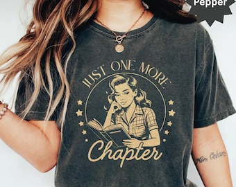 May include: A dark gray t-shirt with a vintage-style graphic of a woman reading a book. The graphic says "Just One More Chapter" in a gold font. The shirt has a distressed look and is made of a soft, comfortable material.