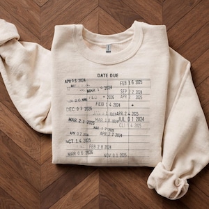 May include: Cream-colored sweatshirt with a printed library card design. The text "DATE DUE" is at the top, with various dates and numbers resembling a library card. The sweatshirt is folded neatly on a wooden surface.