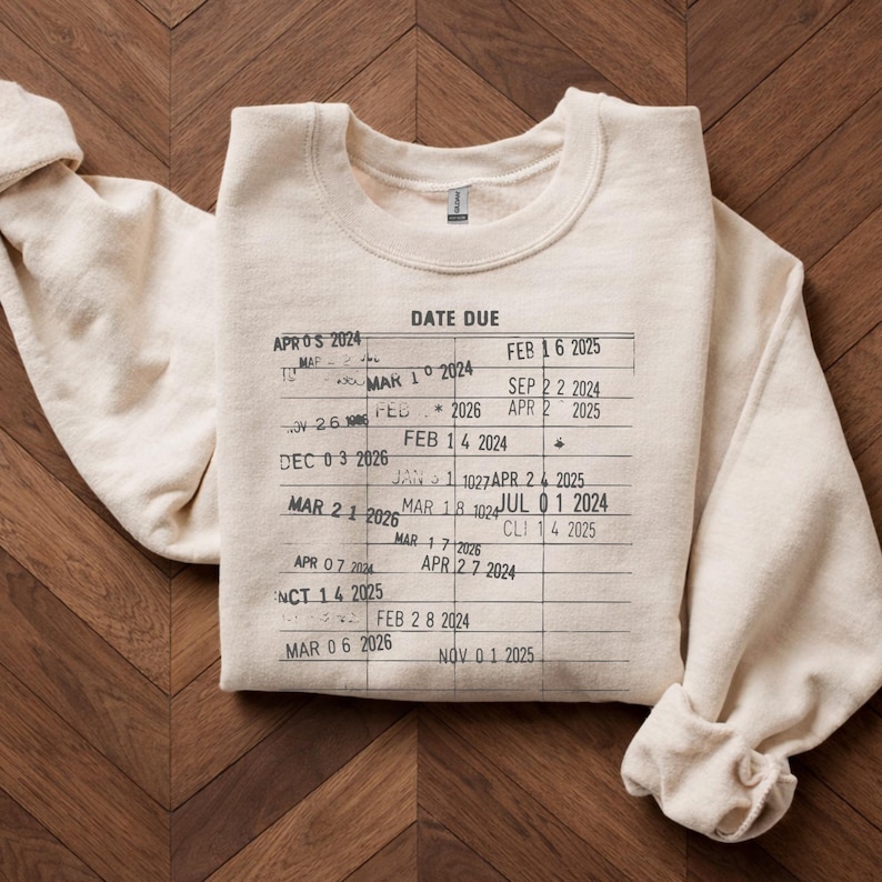 May include: Cream-colored sweatshirt with a printed library card design. The text "DATE DUE" is at the top, with various dates and numbers resembling a library card. The sweatshirt is folded neatly on a wooden surface.