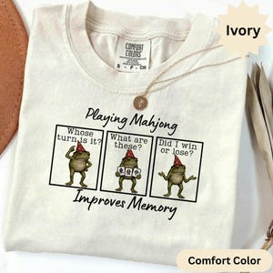 Könnte beinhalten: Ein elfenbeinfarbenes T-Shirt mit einem dreiteiligen Comic-Strip eines Frosches mit rotem Partyhut. Der Text lautet Playing Mahjong, Whose turn is it?, What are these?, Did I win or lose? und Improves Memory. Eine goldene Halskette liegt auf dem Kragen.
