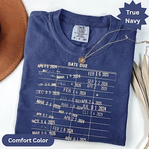 May include: True navy Comfort Colors t-shirt with a vintage-style graphic. The shirt features a list of dates and the words "DATE DUE". A gold necklace with a circular pendant is draped on the shirt.