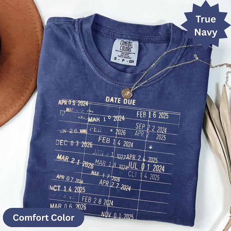 May include: True navy Comfort Colors t-shirt with a vintage-style graphic. The shirt features a list of dates and the words "DATE DUE". A gold necklace with a circular pendant is draped on the shirt.