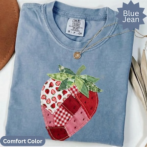 Puede incluir: Una camiseta azul Comfort Colors con un gran diseño de fresa de patchwork. La fresa presenta telas rojas, rosas y blancas, incluido un patrón de cuadros. Un collar de oro con un colgante descansa sobre la camisa. La camisa está etiquetada como "Blue Jean".