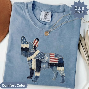 May include: A light blue Comfort Colors t-shirt with a denim patchwork dog design featuring American flag elements. The shirt has a round neckline and short sleeves. A gold necklace with a pendant is draped on the shirt. The words "Blue Jean" and "Comfort Color" are in the image.