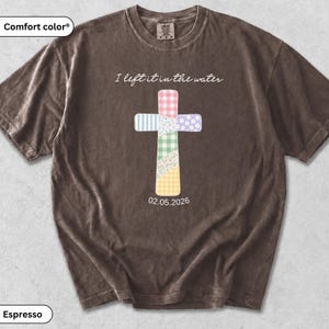 Könnte beinhalten: Ein espressofarbenes Kurzarm-T-Shirt mit einer zentralen Kreuzgrafik, die aus verschiedenen pastellfarbenen, gemusterten Flicken besteht. Der Text "I left it in the water" steht über dem Kreuz und das Datum "02.05.2026" ist darunter aufgedruckt.