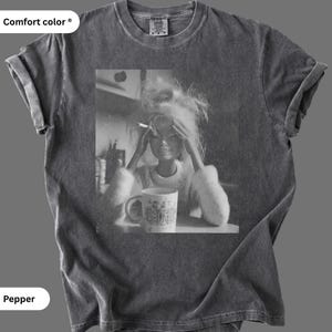 Könnte beinhalten: Ein anthrazitfarbenes Kurzarm-T-Shirt mit Vintage-Waschung. In der Mitte befindet sich eine schwarz-weiße Grafik einer Puppe mit zerzaustem Haar, die eine Zigarette hält und ihre Hände neben einer Tasse mit der Aufschrift GET FOC am Kopf hält.