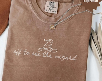 Op de afbeelding: Espresso-kleurig Comfort Colors T-shirt met de tekst "off to see the wizard" en een witte tovenaars hoed afbeelding. Een zilveren ketting met een hanger ligt op het shirt.
