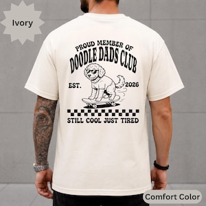 May include: An ivory t-shirt featuring a black graphic on the back. The design shows a dog wearing sunglasses on a skateboard with the text: PROUD MEMBER OF DOODLE DADS CLUB, EST. 2026, and STILL COOL JUST TIRED, along with a checkered pattern.