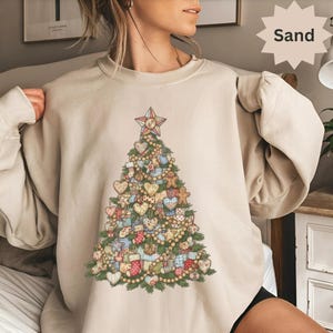Peut inclure: Un sweat-shirt couleur sable avec un motif de sapin de Noël détaillé. L'arbre est orné de cœurs, de bonhommes en pain d'épices, de cadeaux emballés et d'une étoile au sommet. Le motif incorpore diverses couleurs et motifs, créant une esthétique festive et chaleureuse.